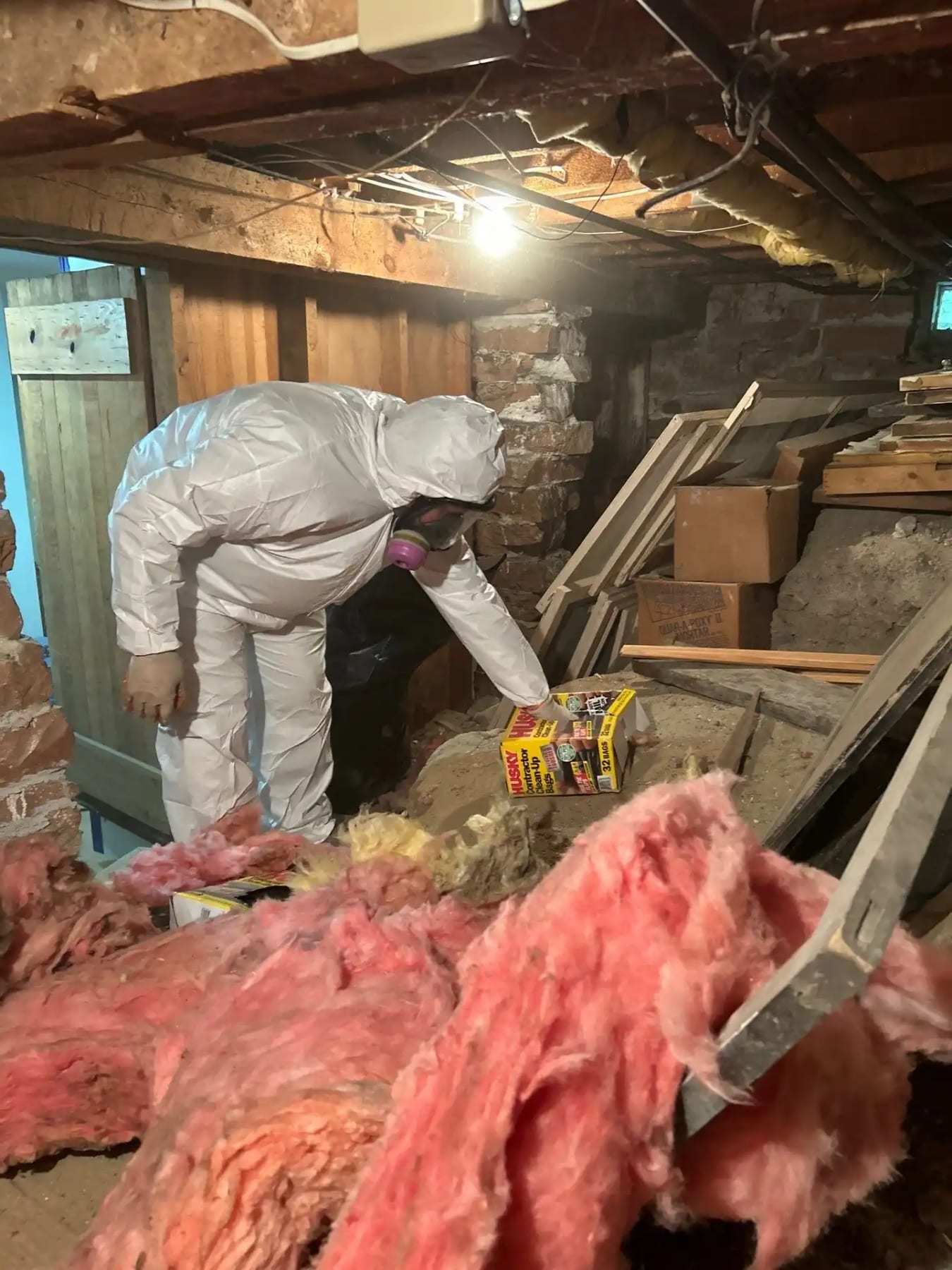 Technician in a protective suit removing wet insulation in a stone-walled crawlspace